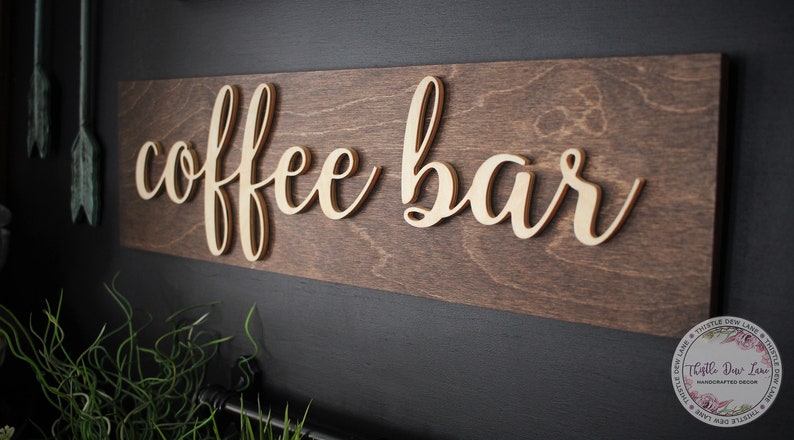 Pu&ograve; includere: Un cartello in legno con le parole "coffee bar" in un font corsivo. Il cartello &egrave; dipinto di colore marrone scuro e le lettere sono di colore marrone chiaro.