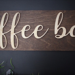 Pu&ograve; includere: Un cartello in legno con le parole "coffee bar" in corsivo, ritagliate nel legno. Il cartello &egrave; marrone e le lettere sono di un colore legno chiaro.