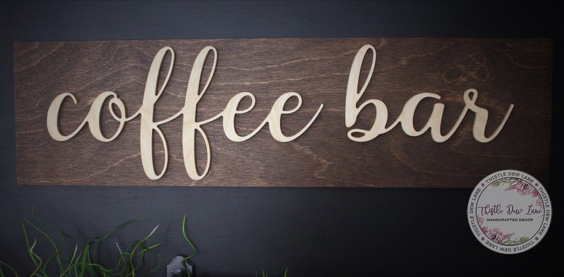 Pu&ograve; includere: Un cartello in legno con le parole "coffee bar" in corsivo, ritagliate nel legno. Il cartello &egrave; marrone e le lettere sono di un colore legno chiaro.