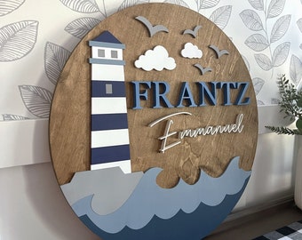 May include: A wooden round sign with a blue and white lighthouse, white clouds, and gray seagulls. The sign also has the name "FRANTZ Emmanuel" in blue and white lettering.