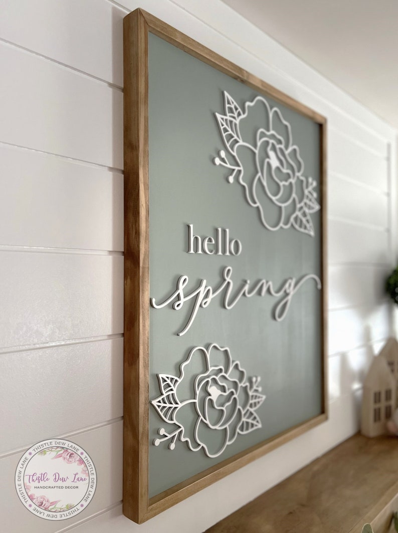 May include: A wooden framed sign with a sage green background and white cutout flowers and the text "hello spring".