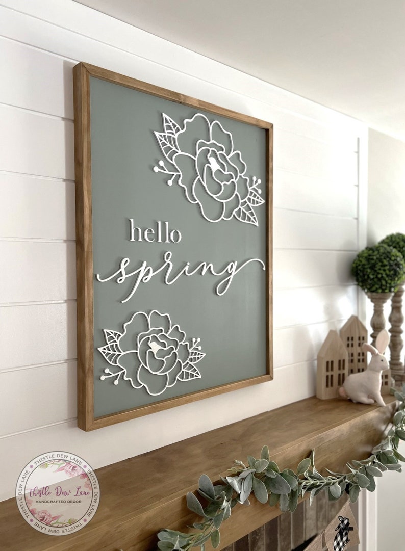 May include: A wooden sign with a sage green background and white lettering that reads "hello spring". The sign is decorated with two white paper flowers.