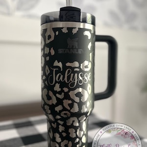 May include: A black stainless steel travel mug with a leopard print design. The mug has a black handle and a clear lid with a straw. The mug is personalized with the name "Jalyss" and the Stanley logo. The mug is sitting on a black and white checkered surface.