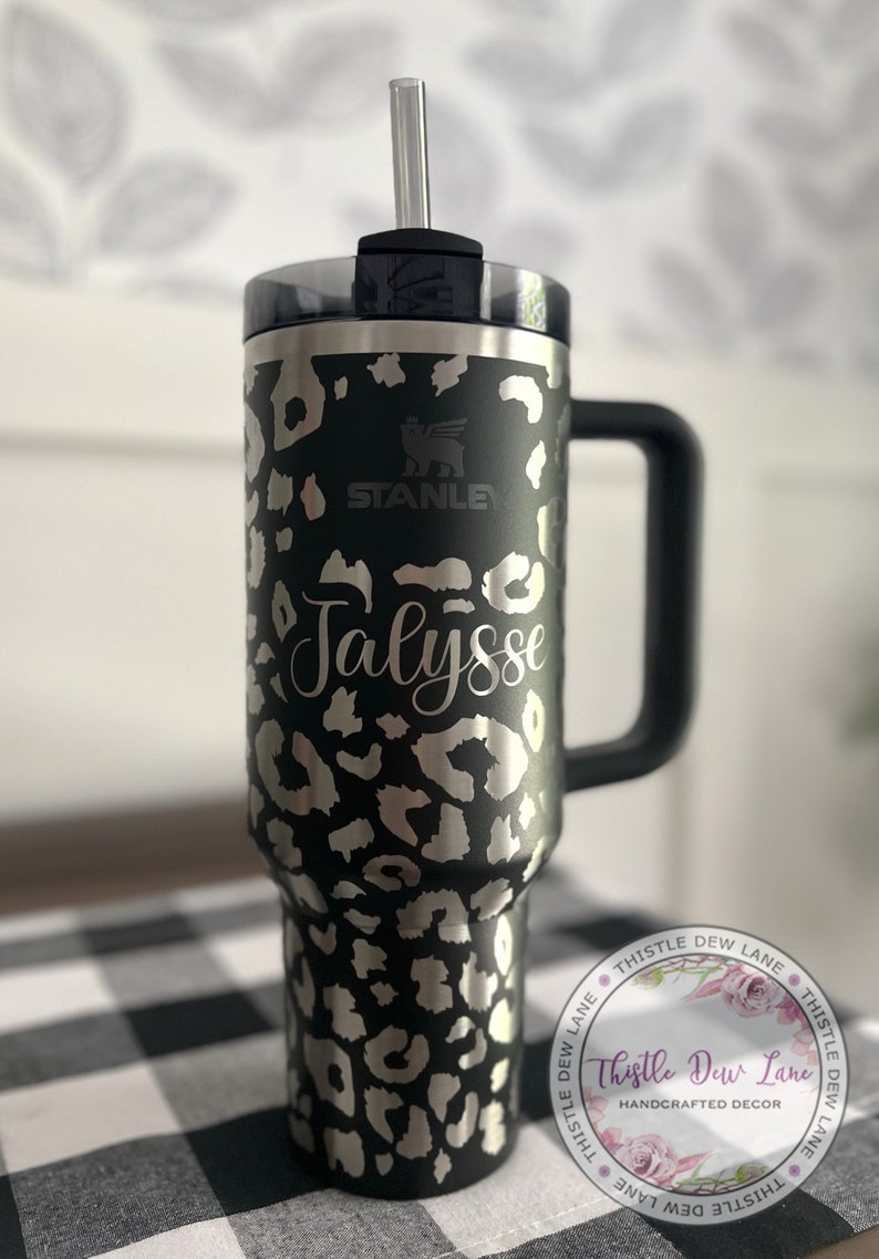 May include: A black stainless steel travel mug with a leopard print design. The mug has a black handle and a clear lid with a straw. The mug is personalized with the name "Jalyss" and the Stanley logo. The mug is sitting on a black and white checkered surface.