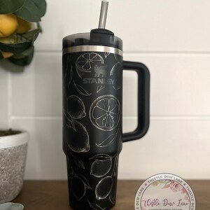 Pu&ograve; includere: Tazzina da viaggio in acciaio inossidabile nero con manico nero e coperchio trasparente. La tazza ha un design a limone argentato e il logo Stanley. La tazza &egrave; su una superficie di legno con un adesivo che dice "Thistle Dew Lane Handcrafted Decor".