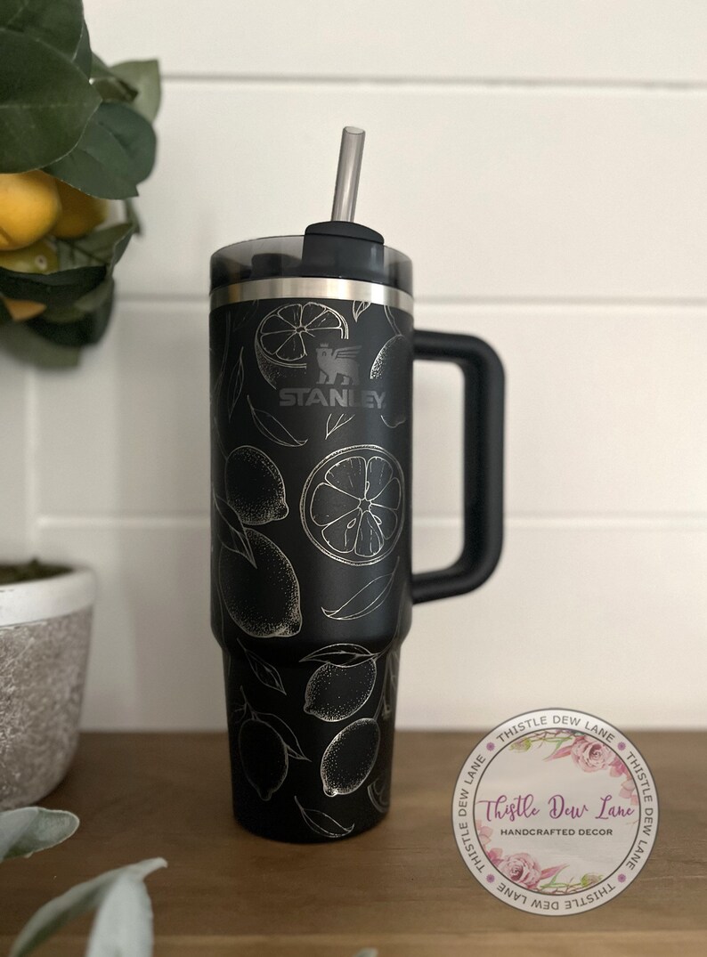 Pu&ograve; includere: Tazzina da viaggio in acciaio inossidabile nero con manico nero e coperchio trasparente. La tazza ha un design a limone argentato e il logo Stanley. La tazza &egrave; su una superficie di legno con un adesivo che dice "Thistle Dew Lane Handcrafted Decor".