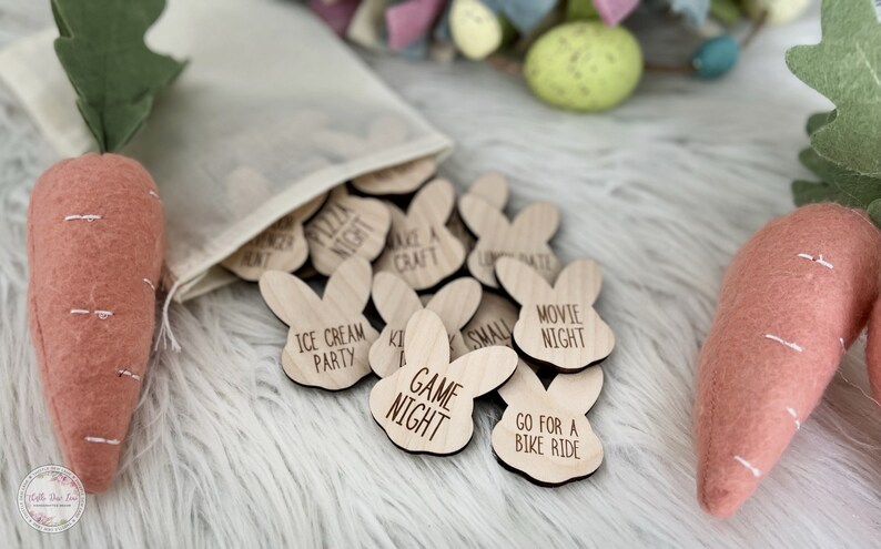 K&ouml;nnte beinhalten: Ein Satz aus Holz-Hasen-Spielsteinen mit verschiedenen Aktivit&auml;ten, die darauf geschrieben sind, wie "Spielabend", "Filmabend" und "Fahrrad fahren". Die Spielsteine befinden sich in einem wei&szlig;en Kordelbeutel und sind von Kunstkarotten und Ostereiern umgeben.