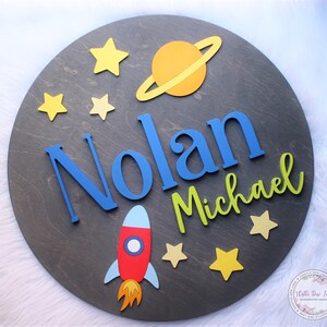 May include: A round wooden sign with a space theme. The sign features a rocket, a planet with rings, stars, and the names Nolan Michael in blue and green letters.