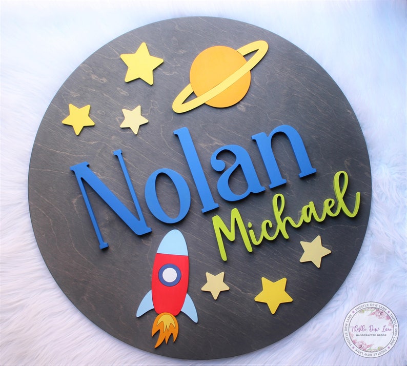 May include: A round wooden sign with a space theme. The sign features a rocket, a planet with rings, stars, and the names Nolan Michael in blue and green letters.