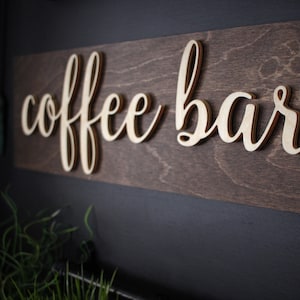 Pu&ograve; includere: Un cartello in legno con la scritta "coffee bar" in lettere dorate. Il cartello &egrave; montato su una parete scura.