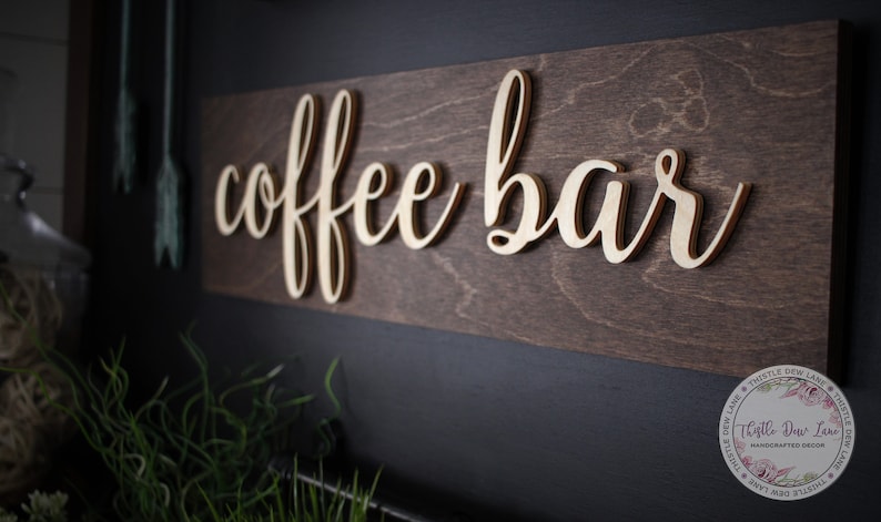 Pu&ograve; includere: Un cartello in legno con la scritta "coffee bar" in lettere dorate. Il cartello &egrave; montato su una parete scura.