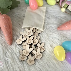 K&ouml;nnte beinhalten: Ein Set aus h&ouml;lzernen, bunnyf&ouml;rmigen Osterei-Spielsteinen mit verschiedenen Aktivit&auml;ten, die darauf geschrieben sind, wie "Filmabend", "Pizzaabend", "Etwas pflanzen" und "Drau&szlig;en spielen". Die Steine befinden sich in einem wei&szlig;en Kordelbeutel und sind von pastellfarbenen Ostereiern umgeben.