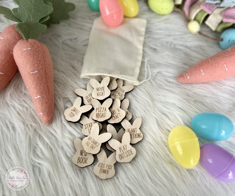K&ouml;nnte beinhalten: Ein Set aus h&ouml;lzernen, bunnyf&ouml;rmigen Osterei-Spielsteinen mit verschiedenen Aktivit&auml;ten, die darauf geschrieben sind, wie "Filmabend", "Pizzaabend", "Etwas pflanzen" und "Drau&szlig;en spielen". Die Steine befinden sich in einem wei&szlig;en Kordelbeutel und sind von pastellfarbenen Ostereiern umgeben.