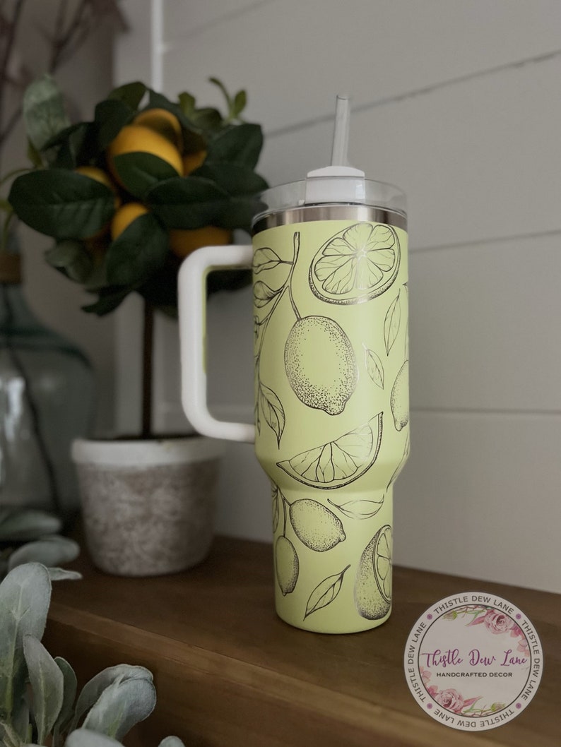 Pu&ograve; includere: Un bicchiere in acciaio inossidabile verde chiaro con un design di limone bianco e nero. Il bicchiere ha un coperchio e un manico. Il bicchiere &egrave; appoggiato su una superficie di legno. Il testo sull'adesivo recita "Thistle Dew Lane Handmade Decor".