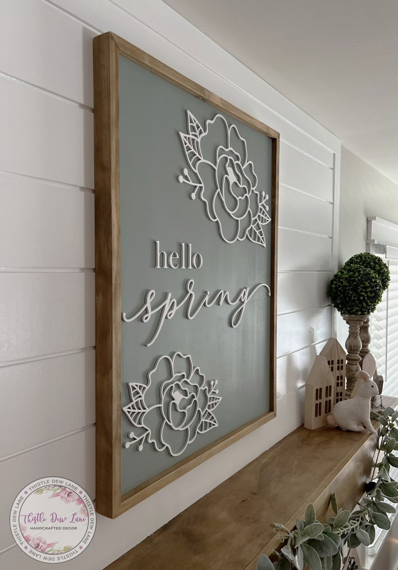 May include: A wooden sign with a light gray background and a white wooden frame. The sign features the text "hello spring" in white script lettering and two white floral designs.
