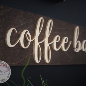 Pu&ograve; includere: Un cartello in legno con le parole "coffee bar" in un font corsivo. Le lettere sono in rilievo e sovrapposte, creando un effetto 3D. Il cartello &egrave; dipinto di un colore marrone scuro e ha un aspetto rustico.