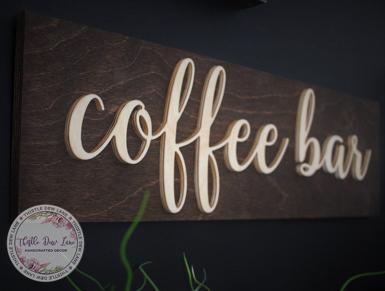 Pu&ograve; includere: Un cartello in legno con le parole "coffee bar" in un font corsivo. Le lettere sono in rilievo e sovrapposte, creando un effetto 3D. Il cartello &egrave; dipinto di un colore marrone scuro e ha un aspetto rustico.