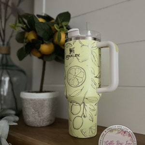 Pu&ograve; includere: Un bicchiere in acciaio inossidabile con un design a stampa di limone. Il bicchiere ha un coperchio bianco e un manico bianco. Il bicchiere &egrave; appoggiato su uno scaffale di legno con una pianta in vaso sullo sfondo. Il bicchiere ha il logo Stanley e le parole "Thistle Dew Lane" su di esso.