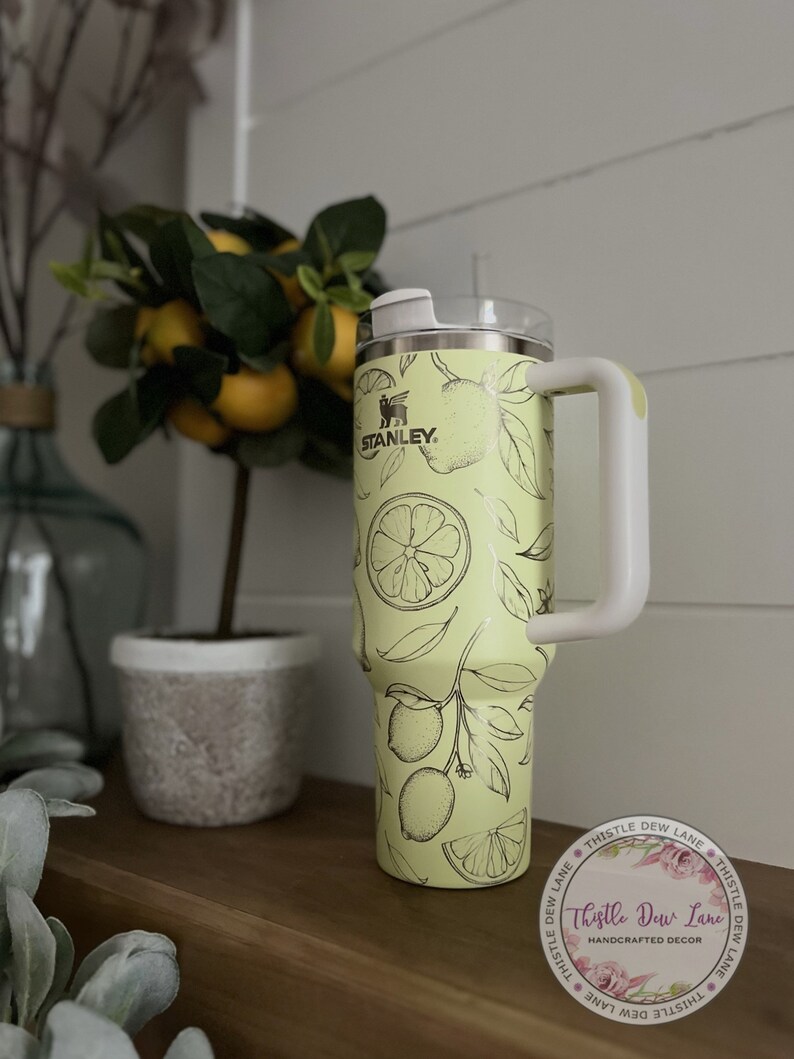 Pu&ograve; includere: Un bicchiere in acciaio inossidabile con un design a stampa di limone. Il bicchiere ha un coperchio bianco e un manico bianco. Il bicchiere &egrave; appoggiato su uno scaffale di legno con una pianta in vaso sullo sfondo. Il bicchiere ha il logo Stanley e le parole "Thistle Dew Lane" su di esso.