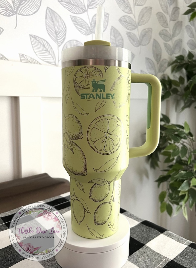 Pu&ograve; includere: Una tazza da viaggio Stanley verde con un design a stampa di limone. La tazza ha un manico e un coperchio con un foro per la cannuccia. La tazza &egrave; appoggiata su una superficie bianca con un motivo a scacchi bianco e nero.