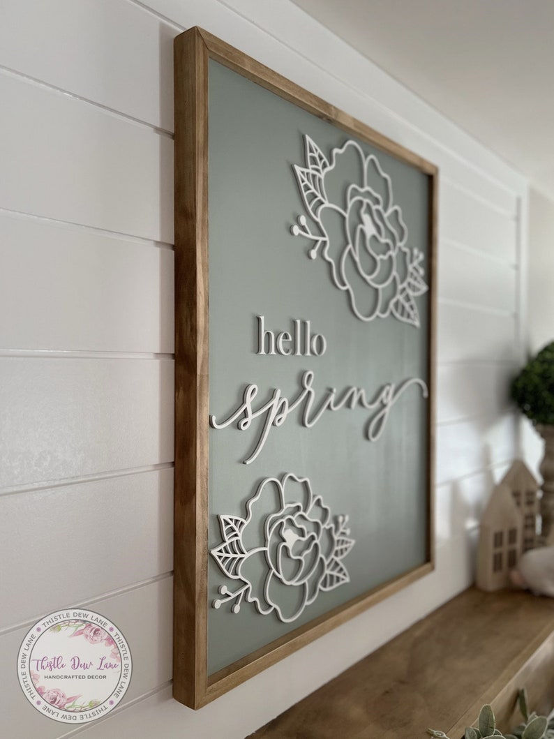 May include: A wooden framed sign with a sage green background and white cutout flowers and the text "hello spring".