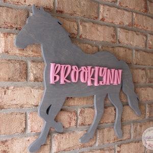May include: A gray wooden horse silhouette with the name "BROOKLYNN" in pink letters. The horse is mounted on a brick wall.