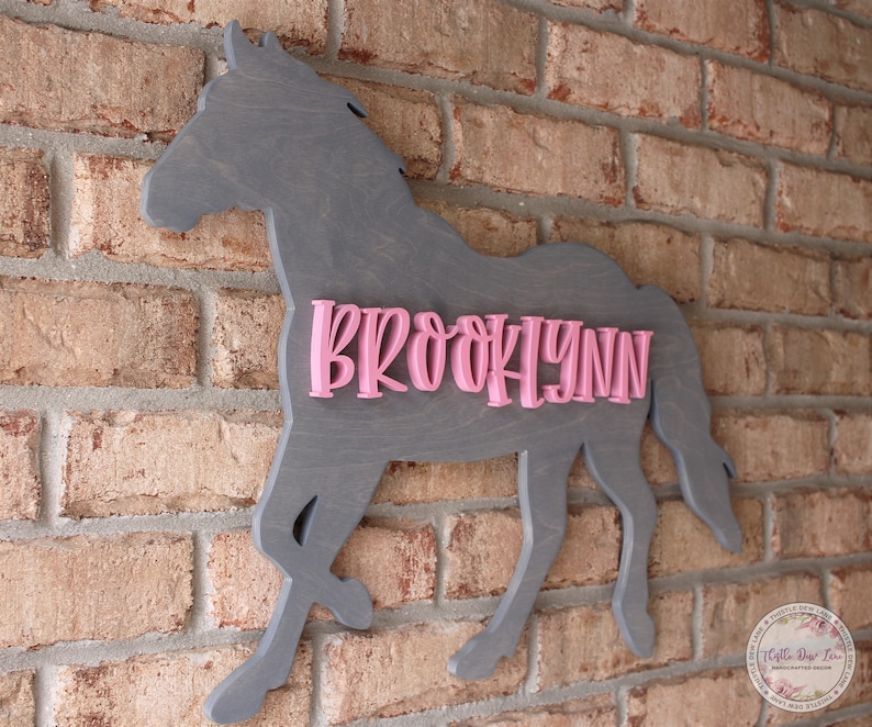 May include: A gray wooden horse silhouette with the name "BROOKLYNN" in pink letters. The horse is mounted on a brick wall.