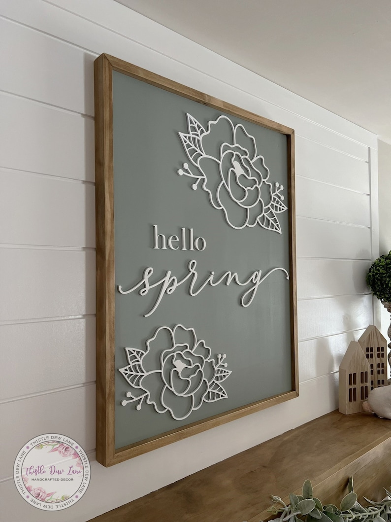 May include: A wooden sign with a light green background and a white wooden frame. The sign says "hello spring" in white script lettering with two white floral designs on either side of the text.
