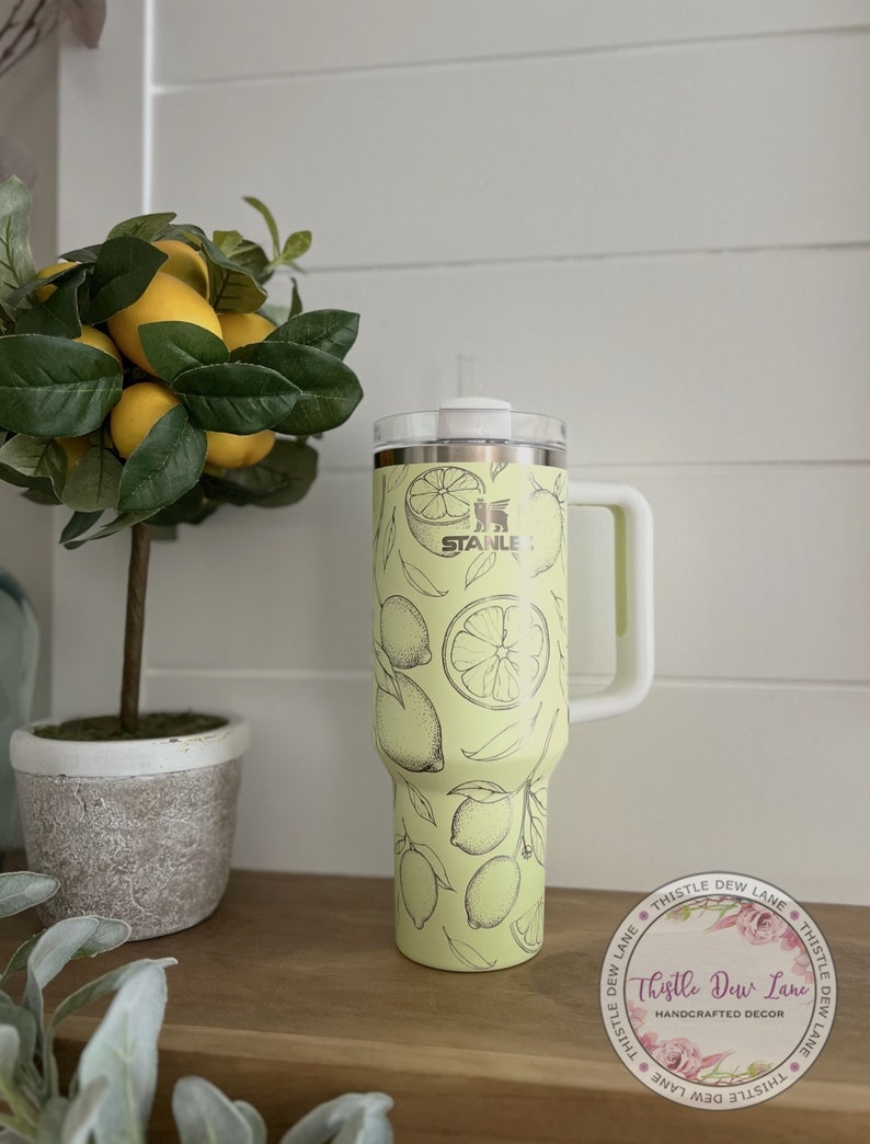 Pu&ograve; includere: Un bicchiere termico in acciaio inossidabile giallo con un design a limone. Il bicchiere ha un coperchio e un manico. Il bicchiere &egrave; appoggiato su una superficie di legno. Il testo "STANLEY" &egrave; visibile sul bicchiere. Il testo "Thistle Dew Lane HANDCRAFTED DECOR" &egrave; visibile su un adesivo.