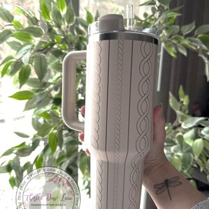 May include: A light pink stainless steel tumbler with a rope design and a lid. The tumbler has a handle and a straw. The tumbler is being held by a person with a dragonfly tattoo on their wrist. The tumbler has the text "Thistle Dew Lane" on it.