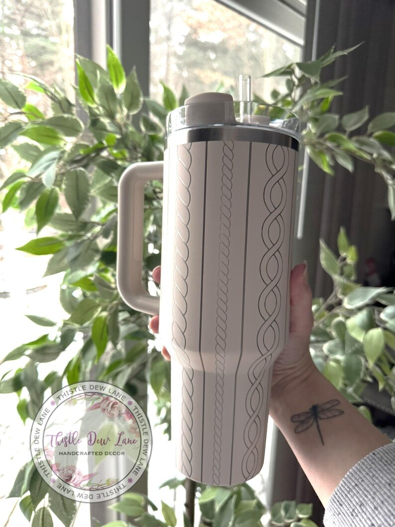 May include: A light pink stainless steel tumbler with a rope design and a lid. The tumbler has a handle and a straw. The tumbler is being held by a person with a dragonfly tattoo on their wrist. The tumbler has the text "Thistle Dew Lane" on it.