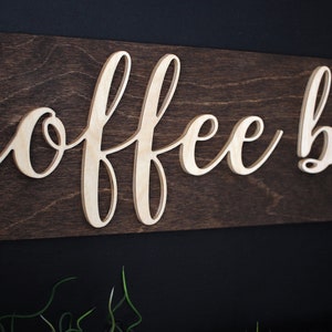 Pu&ograve; includere: Un cartello in legno con le parole "coffee bar" in lettere bianche in rilievo su uno sfondo marrone scuro.