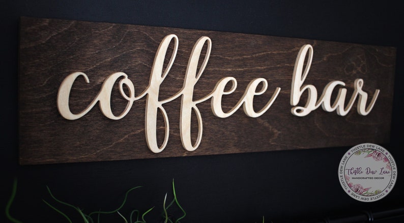 Pu&ograve; includere: Un cartello in legno con le parole "coffee bar" in lettere bianche in rilievo su uno sfondo marrone scuro.