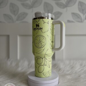 Pu&ograve; includere: Una tazza da viaggio in acciaio inossidabile gialla con un coperchio e un manico bianchi. La tazza ha un design a stampa di limone con contorni neri e la parola "STANLEY" in lettere nere.