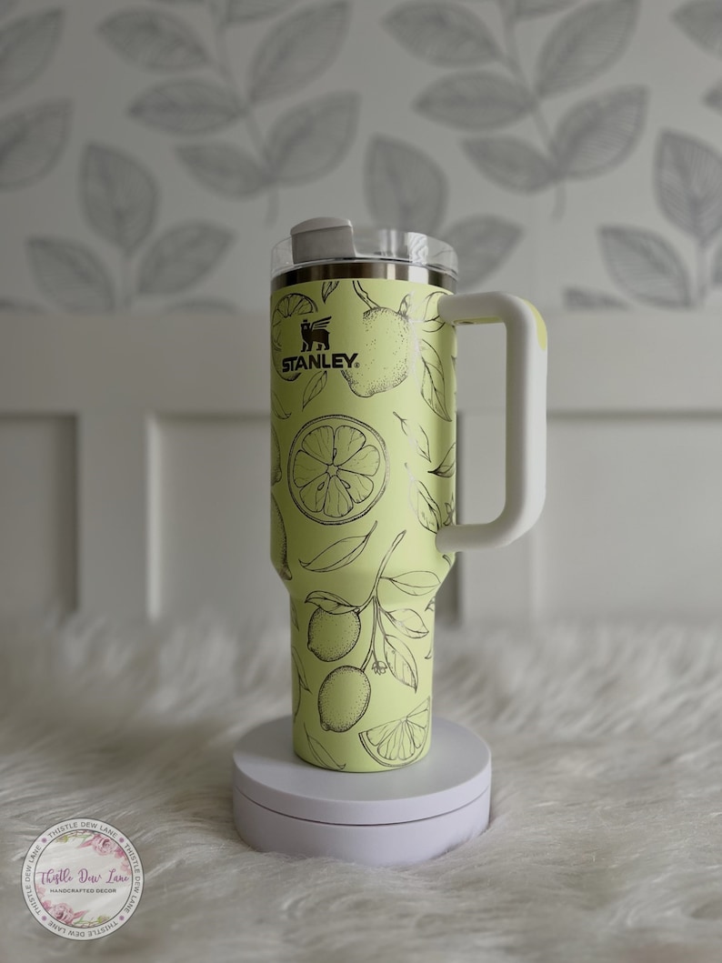 Pu&ograve; includere: Una tazza da viaggio in acciaio inossidabile gialla con un coperchio e un manico bianchi. La tazza ha un design a stampa di limone con contorni neri e la parola "STANLEY" in lettere nere.