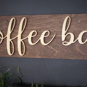 Pu&ograve; includere: Un cartello in legno con le parole "coffee bar" in lettere dorate. Il cartello &egrave; montato su una parete marrone scuro.