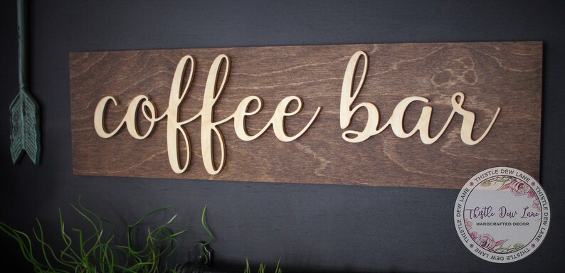 Pu&ograve; includere: Un cartello in legno con le parole "coffee bar" in lettere dorate. Il cartello &egrave; montato su una parete marrone scuro.