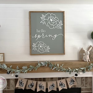 May include: A white wooden sign with the text "hello spring" in black script font and a floral design. The sign is hanging above a fireplace mantel decorated with a garland of eucalyptus leaves and a banner of black and white checkered bunny flags.