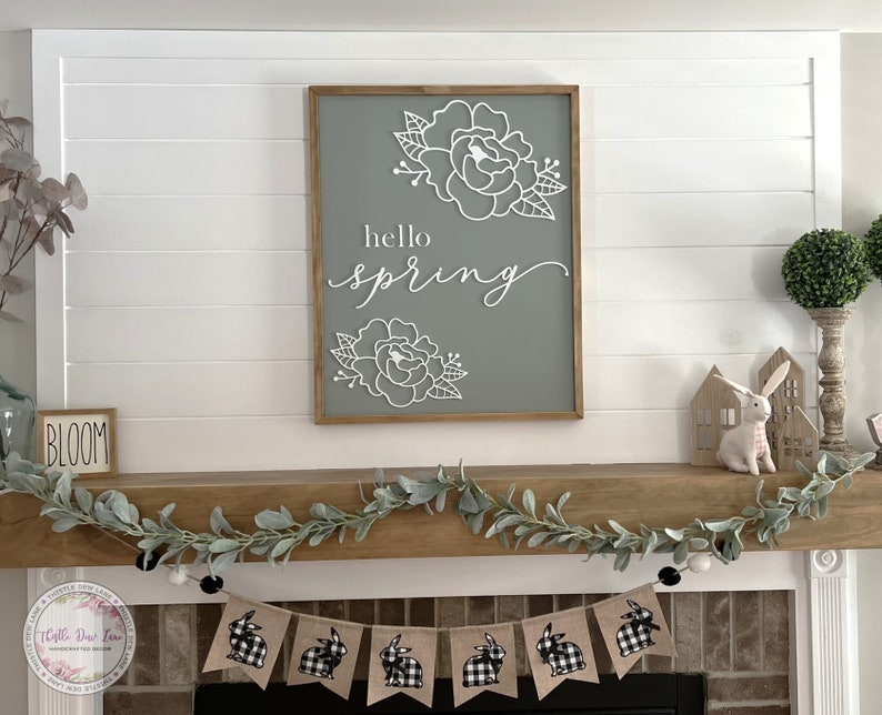 May include: A white wooden sign with the text "hello spring" in black script font and a floral design. The sign is hanging above a fireplace mantel decorated with a garland of eucalyptus leaves and a banner of black and white checkered bunny flags.