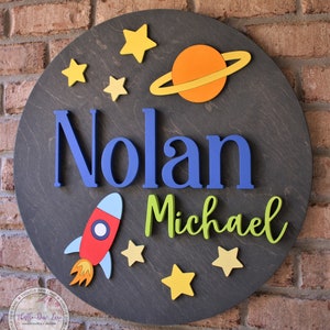 May include: A round wooden sign with a space theme. The sign features the names "Nolan Michael" in blue and green letters, a red and blue rocket, a yellow and orange planet with rings, and yellow stars.