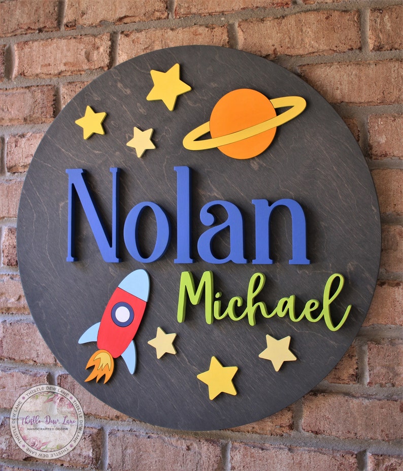 May include: A round wooden sign with a space theme. The sign features the names "Nolan Michael" in blue and green letters, a red and blue rocket, a yellow and orange planet with rings, and yellow stars.