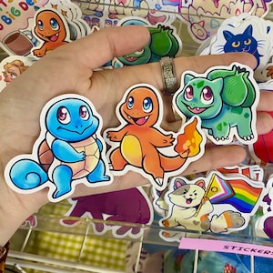 May include: Four colorful cartoon stickers featuring characters from the Pokemon franchise. The stickers are on a hand, and the characters are Squirtle, Charmander, Bulbasaur, and a cat with a rainbow flag.