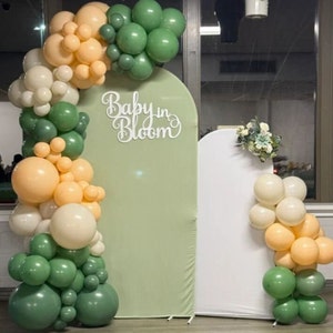 May include: A green and white balloon arch with the words "Baby in Bloom" in white letters. The arch is in front of a green backdrop and a white backdrop.