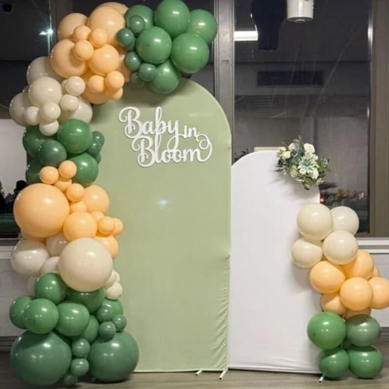 May include: A green and white balloon arch with the words "Baby in Bloom" in white letters. The arch is in front of a green backdrop and a white backdrop.