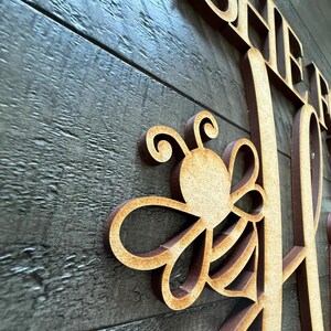 May include: Wooden sign with the word "BEE" and a stylized bee design. The sign is made of light brown wood and mounted on a dark gray wooden background. The bee has a circular body and curved wings.