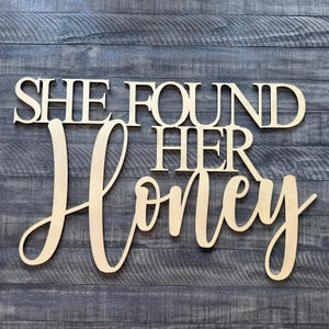 May include: Wooden sign with the phrase "SHE FOUND HER Honey" in a cursive font. The sign is made of light-colored wood and is mounted on a dark gray wooden background. The sign is suitable for home decor.