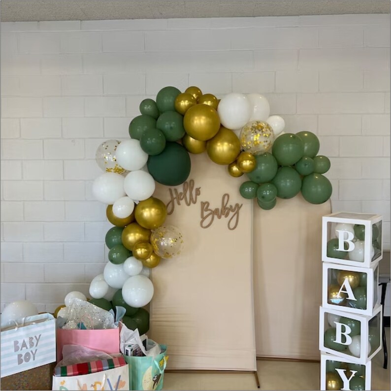May include: A green, gold, and white balloon arch with the words "Hello Baby" in brown script. There are white gift bags in the foreground and a stack of white boxes with the letters "BABY" in black.