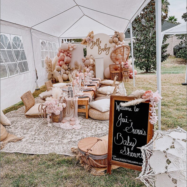 May include: A baby shower celebration with a boho theme. The setup includes a white tent, a backdrop with balloons and greenery, a table with a floral centerpiece, and a chalkboard sign that says "Welcome to Jai + Sura's Baby Shower".