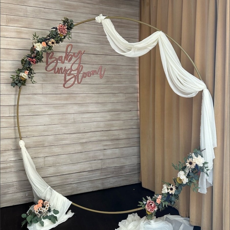 Op de afbeelding: Een gouden metalen hoepelboog versierd met witte stof en roze en groene bloemstukken. De boog is versierd met de tekst "Baby and Bloom".