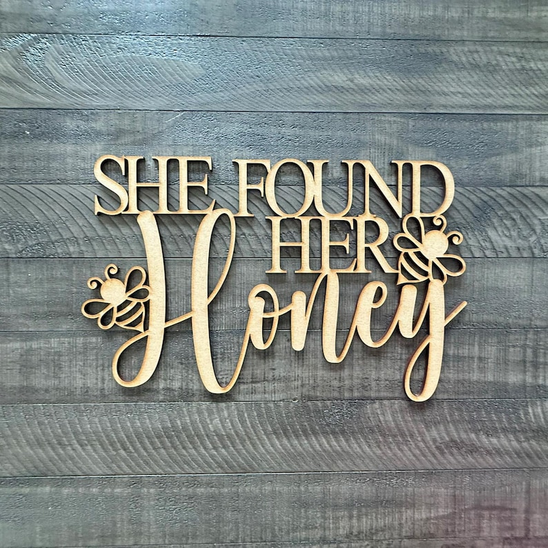 May include: Wooden sign with the phrase "SHE FOUND HER Honey" in a cursive font. The word "Honey" has a bee design on each side. The sign is made of light-colored wood and is set against a gray wood-grain background.